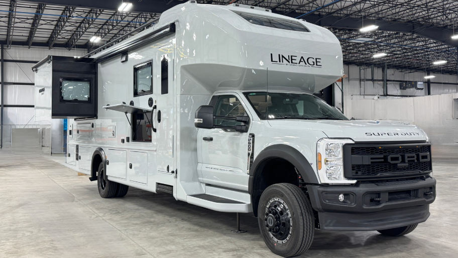 Image of a modern and sleek white motorhome built on a Ford Super Duty chassis, branded as 'Lineage.' The motorhome features multiple compartments, slide-outs, and a raised sleeping area above the cab. The vehicle is parked in a spacious indoor facility with high ceilings and industrial lighting.