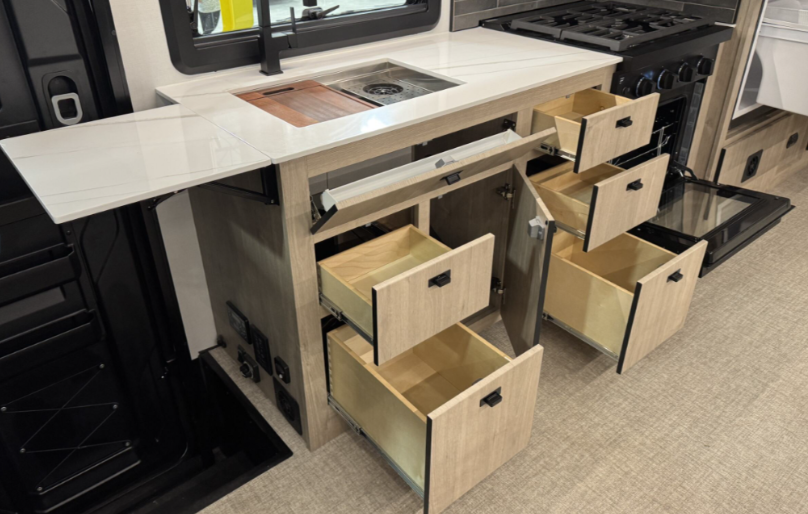 Close-up of a motorhome kitchen area showcasing its efficient storage solutions. The light wood cabinetry includes multiple pull-out drawers and cabinets, providing ample space for organization. A stainless steel sink with an integrated cutting board and a fold-out countertop extension maximize functionality. Adjacent to the sink is a black stovetop and oven, complementing the modern and practical design.