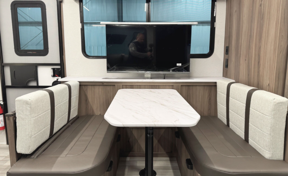 An RV dining area featuring a booth-style setup with a white marble-patterned table flanked by cushioned benches. The benches have light-colored fabric backs with brown accents and brown leather seats. Above the dining table, a flat-screen TV is mounted against a woodgrain panel, offering entertainment for the space. Large windows behind the TV and on the side provide natural light, enhancing the airy and modern feel. To the left, part of the entry door and a fire extinguisher are visible, adding practicality to the design.