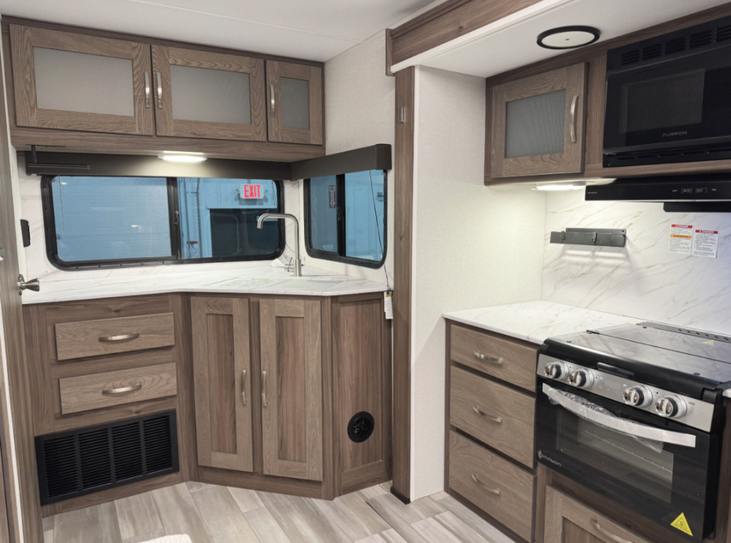 Interior of an RV kitchen showcasing a modern design with woodgrain cabinets and drawers. The space features a corner sink with a stainless steel faucet under a large window, providing natural light. A three-burner stovetop and oven are on the right, with a microwave and additional storage cabinets above. Marble-patterned countertops and backsplash enhance the elegant look, while the lower section includes vented panels and ample drawer space. The layout maximizes functionality and aesthetic appeal in a compact area.