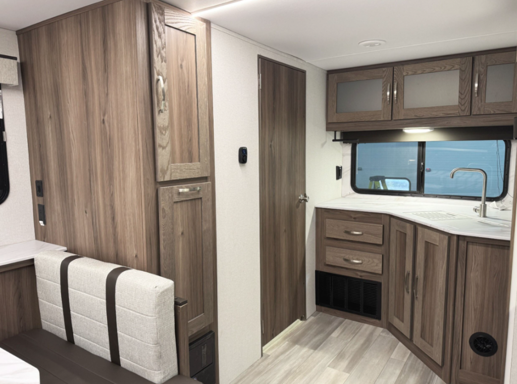 Interior view of an RV kitchen and dining area featuring woodgrain cabinets and drawers, a corner sink with a stainless steel faucet, and a large window above the countertop. The design includes ample storage, with glass-front upper cabinets and concealed lower cabinets. To the left, part of the dining booth is visible, showcasing cushioned seating with a striped pattern. A wooden door in the center leads to another area of the RV, complementing the cohesive woodgrain aesthetic.