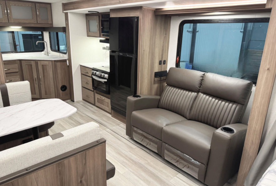 An RV interior showcasing a cozy living area with a focus on functionality. On the right, there’s a dark brown leather loveseat with cupholders integrated into each armrest. Adjacent to the seating area, a large window allows natural light to fill the space. In the background, a sleek kitchen features woodgrain cabinetry, a black refrigerator, a stove with an oven, and a countertop with a stainless steel sink under a corner window. To the left, a booth-style dining area with a white marble-patterned table and cushioned seats adds to the modern, practical design of the space. Light wood flooring ties the entire interior together, creating a warm and inviting atmosphere.