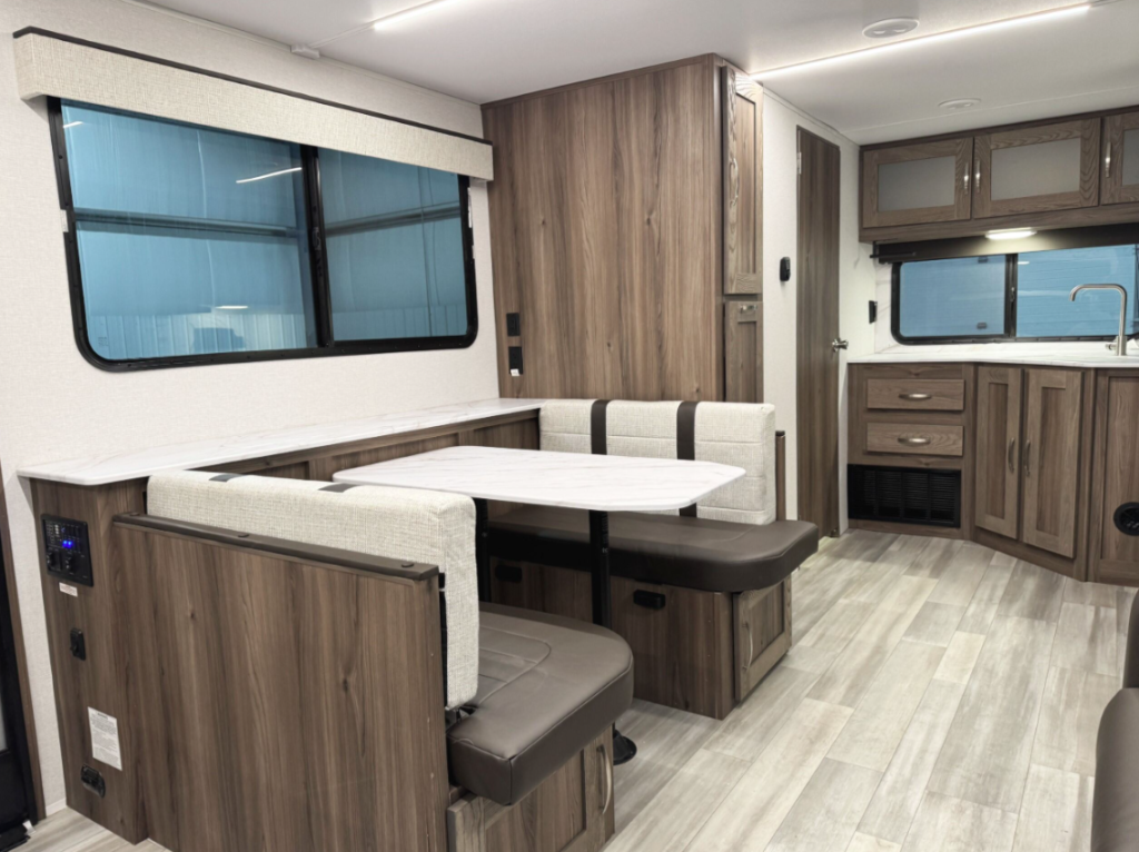 An RV interior with a dining and kitchen area. The dining space features a booth-style setup with a white marble-patterned tabletop and cushioned seats in neutral tones. A large window with a fabric valance provides natural light and an open feel. Adjacent to the dining area, the kitchen showcases woodgrain cabinetry, a corner sink with a stainless steel faucet, and additional storage above and below. The light-toned flooring complements the modern, cohesive design of the space.