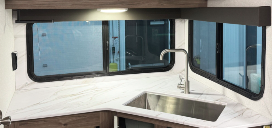 Modern RV kitchen with a marble-look countertop, stainless steel sink, high-arc faucet, and large windows providing ample natural light. The countertop features a wood-tone base that complements the contemporary design.