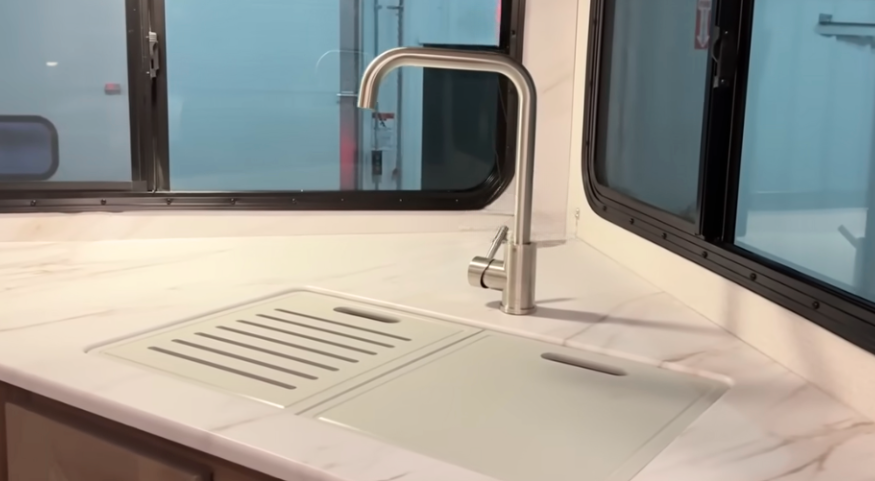 Close-up of a modern RV kitchen counter featuring a marble-look surface with an integrated sink covered by a dual sink cover, including a slotted draining section. A sleek, high-arc stainless steel faucet is positioned in front of large windows for a bright and open feel.