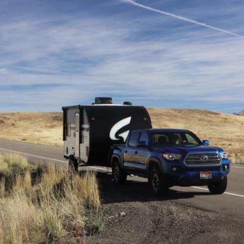 A blue Toyota Tacoma pickup truck towing a black and white travel trailer with a bold "G" logo, parked on the side of a winding road in a vast, open landscape. The terrain features rolling golden hills covered in dry grass under a bright blue sky with wispy clouds and contrails. The remote and scenic backdrop suggests an adventurous road trip or camping excursion in a rural or desert-like setting.